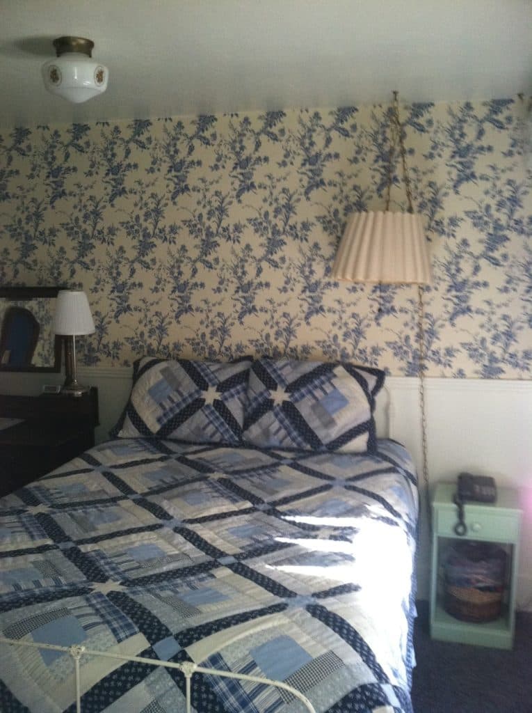 A cozy bedroom featuring a quilted bedspread, floral wallpaper, and vintage decor.