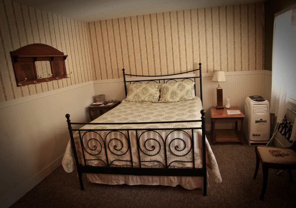 A cozy bedroom featuring a metal bed frame, patterned bedding, a nightstand with a lamp, and a window.