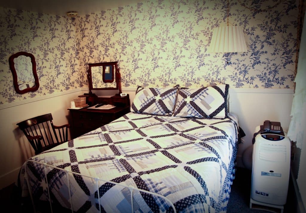 A cozy bedroom with floral wallpaper, a patterned quilted bed, a wooden chair, and a lamp.