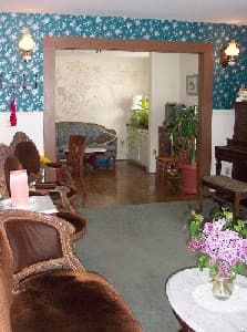 A cozy living room with floral wallpaper, vintage furniture, and a view into another room filled with plants and a piano.