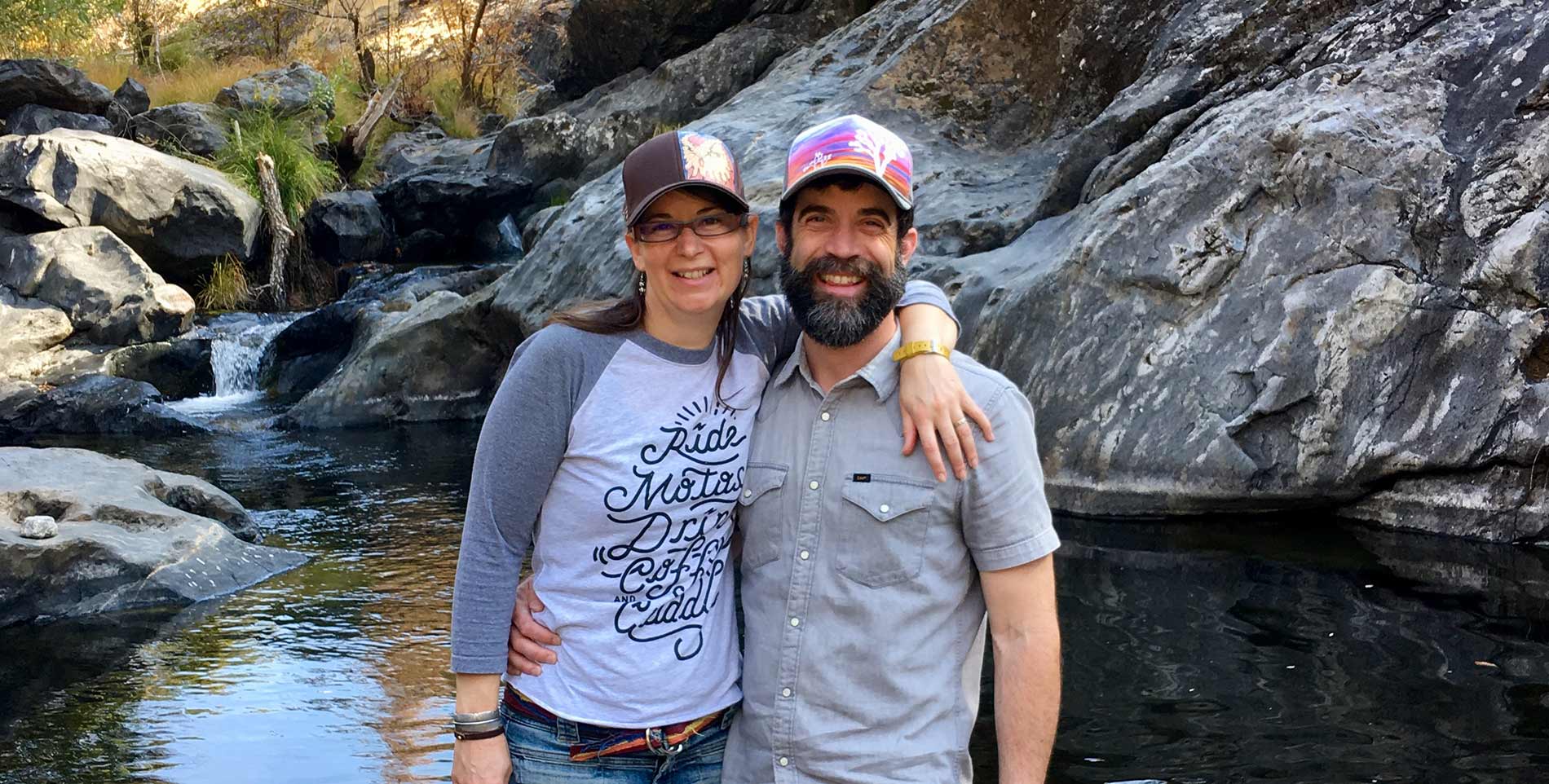 Innkeepers in ballcaps standing in front of a stream, embracing shoulder to shoulder.