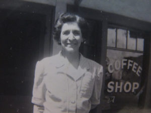 1940's black and white image of original owner of the hotel, Charlotte DeFerrari standing in front of her restaurant window that reads "Coffee Shop."