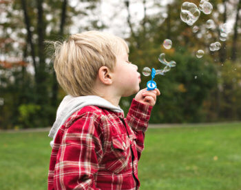 image of towheaded boy with red plaid jacket blowing bubbles on green lawn and trees in the background