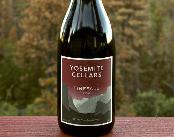 bottle of red wine with burgundy label with shades of gray mountains on label from Yosemite Cellars on red painted table in fall foliage trees in background