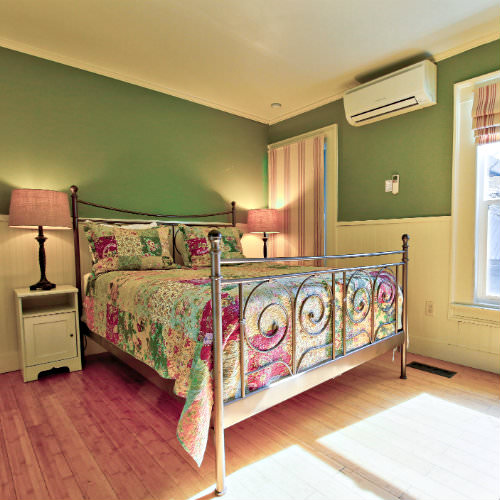 Green guest room with hardwood flooring, double nightstands, two large windows, and multi-colored quilt bedding