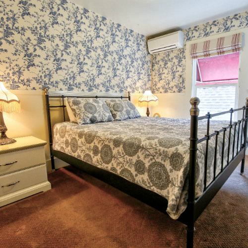 Blue floral wallpapered guest room with brown carpeted floors, grey and white bedding, and double nightstands with reading lamps