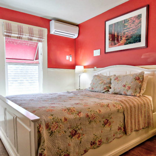 Large white wood bed with white, red and green floral and stripe quilt in room with white beadboard and red painted walls in front of large window with natrual light.