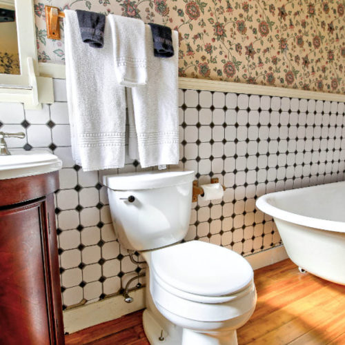 Bathroom with wood vanity, white commode and clawfoot tub with black and white checked wallpaper from the floor to chair rail and complementary beige wallpaper with small floral print.