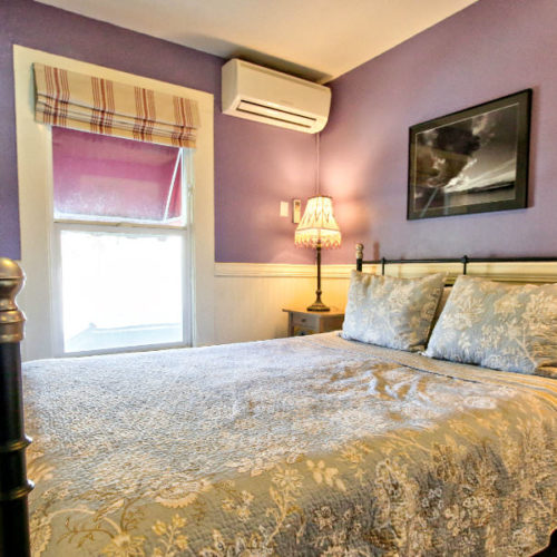 Large bed with cream and lavender quilt in room with white painted beadboard half way up wall, lavender painted walls with lamp on night stand in corner in front of large window with natural light.