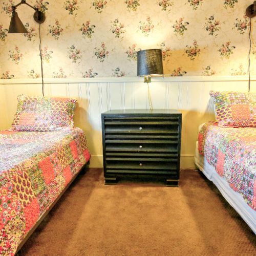 Twin beds covered in pale pink and green matchingquilts with painted wood chest of drawers in between with small lamp in room with traditional cream wallpaper with small florals.