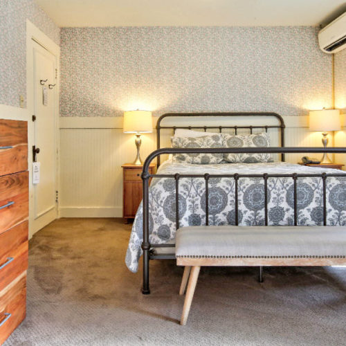 Large black cast iron bed covered in blue and white quilt in carpeted room with covered bench in front, and soft blue colored walls.