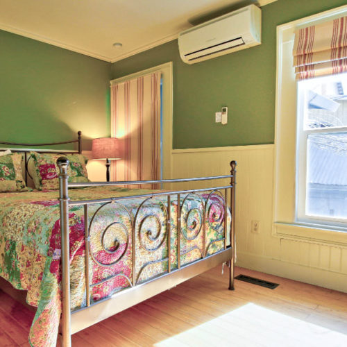 Large cast iron bed covered in green and red patterned quilt with white bead board half way up the wall and corresponding green painted walls with large window letting in natural light.
