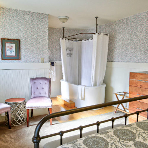 Black cast iron bed with green and white pattered quilt facing large soaking tub with white shower curtain next too a wood chest of drawers and two soft pink tuffted chairs.