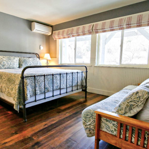 Large cast iron bed with neutral linens on wood floor with soft gray walls and a wood futon with corresponding cover in front of bank of four windows