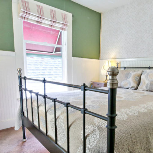 Large cast iron bed covered in green and red patterned quilt with white bead board half way up the wall and corresponding green painted walls with large window letting in natural light.