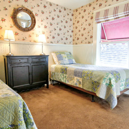 Twin beds covered in pale pink and green matchingquilts with painted wood chest of drawers in between with small lamp in room with traditional cream wallpaper with small florals.