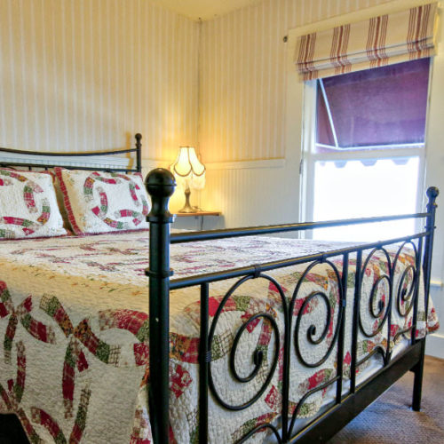 Large black cast iron bed with cream quilt with green and soft pink design in room with cream walls, matching striped window treatment and large window with natural light.