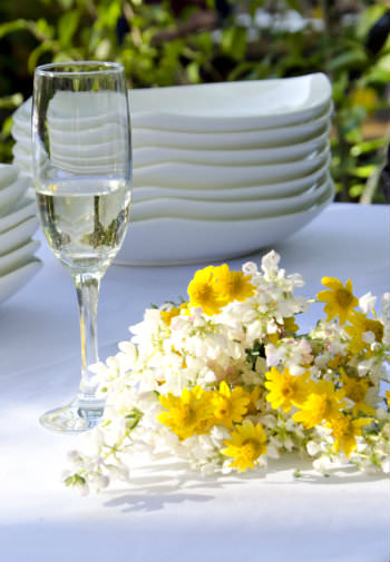 A bouquet of white and yellow flowers by a wineglass a stack of dinner plates all outdoor on a table