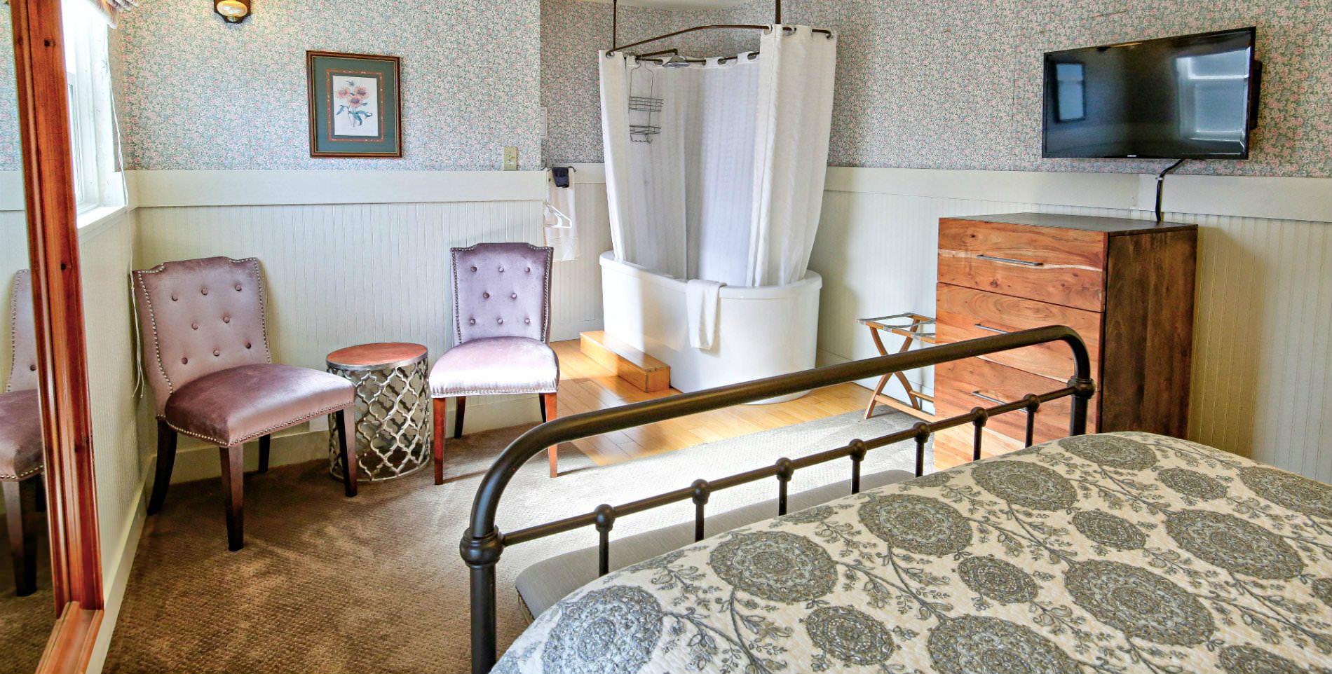 natural light filled guestroom with large black cast iron bed with white soaking tub and shower curtain in the background next to tall medium wood chest of drawers with flat screen tv above and two pale pink apolstered side chairs against wall