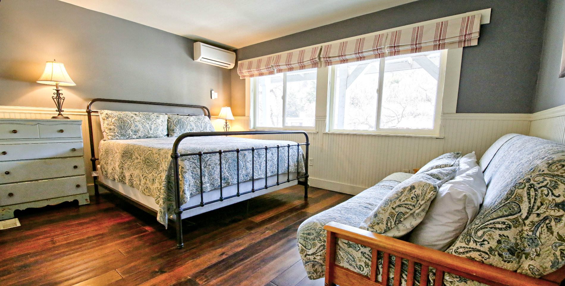 large black cast iron bed with neutral soft gray linens in room with hardwood floors and four windows with lots of natural light and wood futon bed with matching linens